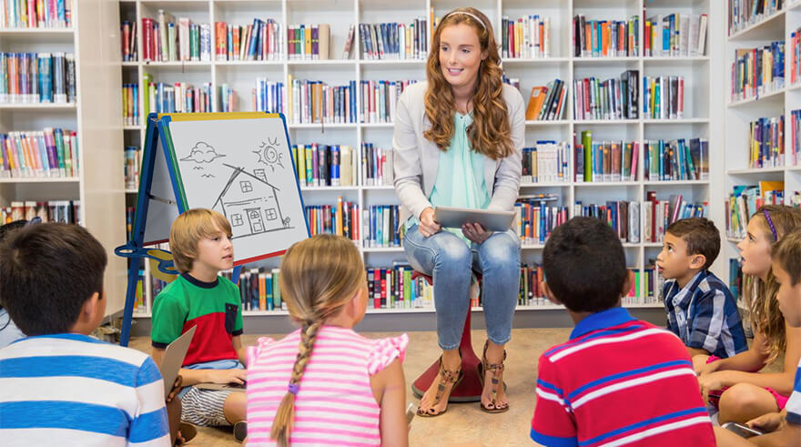 Teacher holding a tablet and sitting on a bench surrounded by children sitting on the floor in a library.