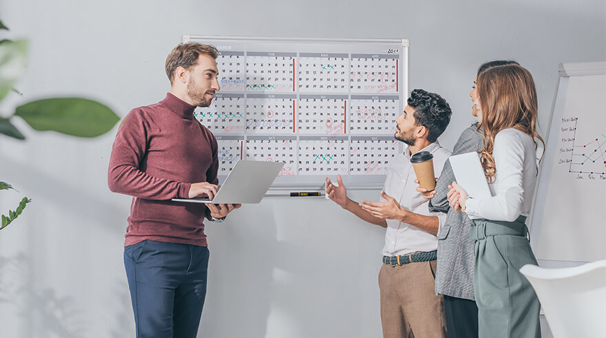 Four employees standing up next to a yearly planner discussing a business plan.